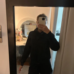 Svart hoodie med huva - En basic svart hoodie med huva och långa ärmar. Tröjan har en klassisk passform och är perfekt för chill dagar. Mjuk insida och ribbade muddar vid ärmslut och nederkant. Enkel och stilren design utan tryck eller logga.