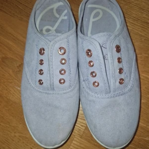 Lila sneakers utan snörning - Snygga lila sneakers i tyg med metallfärgade öljetter och klassisk rund tå. Skorna har en enkel och stilren design utan snören, perfekt för en avslappnad look.