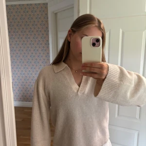 Beige stickad tröja med krage Lindex - Supermjuk och beige stickad tröja från Lindex med breda ärmar och snygg krage. Tröjan har v-ringning och ribbade muddar vid ärmslut och nederkant. Perfekt för lager-på-lager och chill vibes.