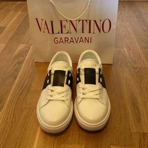 Valentino Garavani Open sneakers vit/svart - Snygga Valentino Garavani Open sneakers i vitt skinn med bred svart läderdetalj över mitten. Klassisk rund tå, vita snören och ikoniska nitar baktill. Perfekta för dig som vill ha en clean men lyxig look.