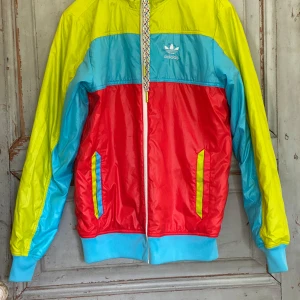 Färgglad Adidas vändbar vindjacka med huva  - Säljer en riktigt färgstark vindjacka från Adidas med huva och dragkedja. Jackan har coola blockfärger i gult, rött och blått, samt ribbade muddar och fickor med kontrastdetaljer. Perfekt för dig som vill sticka ut och gillar retro vibes. Jackan är vändbar!