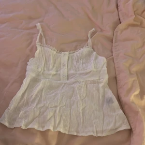 Vit linnetopp med spets och knappar - Supersöt vit linnetopp med tunna axelband, spetsdetalj vid urringningen och tre knappar framtill. Tunt, krinklat bomullstyg som ger en luftig känsla. Perfekt för varma dagar och enkel att styla med jeans eller kjol.