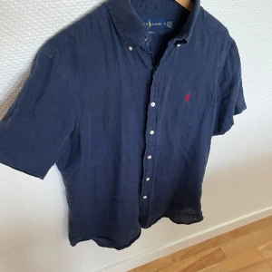 Mörkblå linneskjorta från Ralph Lauren - Snygg mörkblå skjorta från Ralph Lauren i 100% linne med korta ärmar och klassisk button-down krage. Skjortan har vita knappar framtill och den ikoniska röda Polo-loggan broderad på bröstet. Perfekt för en chill och stilren look.