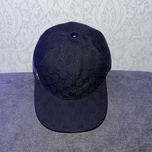 Svart Gucci keps med GG-mönster - Snygg svart keps från Gucci med klassiskt GG-monogram över hela kepsen och ikoniska gröna och röda ränder på sidorna. Justerbar rem baktill för perfekt passform. Tillverkad i bomull och polyester, perfekt för dig som vill sticka ut med lyxig streetstyle.