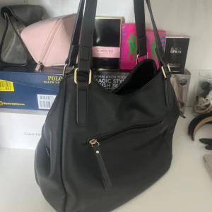 Svart axelväska med gulddetaljer - Stilren svart axelväska i skinnimitation med flera fack och dragkedja framtill. Väskan har breda axelband och snygga guldfärgade metalldetaljer. Perfekt för dig som vill ha plats för allt du behöver under dagen och ändå hålla stilen. Nypris 700