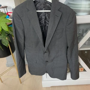 Mörkgrå kavaj Oscar Jacobson 48 - Snygg mörkgrå kavaj från Oscar Jacobson, modell Edmund Blazer. Klassisk passform med två knappar framtill, diskreta fickor och helfodrad insida. Perfekt för dig som vill ha en stilren och tidlös look.