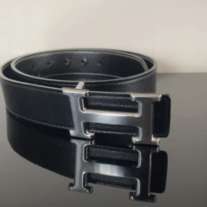 Svart bälte med H-spänne - Snyggt svart bälte med ikoniskt H-format spänne i silverfärgad metall. Bältet är gjort i äkta skinn med en stilren och klassisk design som passar till många outfits. Perfekt accessoar för dig som gillar exklusiva detaljer.