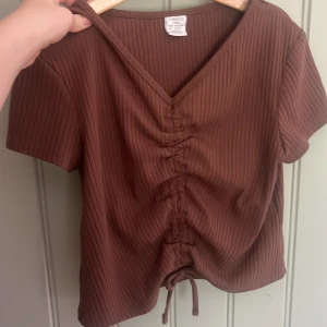 Brun ribbad topp med snörning Lindex - Brun ribbad topp från Lindex med kort ärm och v-ringning. Toppen har snygg snörning framtill som ger en cool rynkad effekt och croppad passform. Perfekt för dig som gillar trendiga detaljer och enkel stil.
