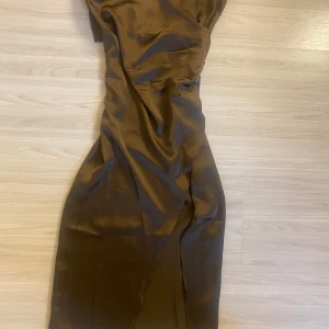 Brun omlottklänning i satin - Snygg brun långklänning i satin med omlott-detalj och smala axelband. Klänningen har en slits framtill och en figurnära passform som ger en elegant siluett. Perfekt för dig som vill sticka ut med en stilren look.