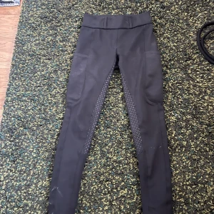 Svarta ridleggings med grepp - Svarta ridleggings med helskodd grepp i sätet och längs insidan av benen. Byxorna har praktiska fickor på sidorna och är tillverkade i stretchigt material av polyester och spandex. Perfekta för ridning och ger bra rörlighet.