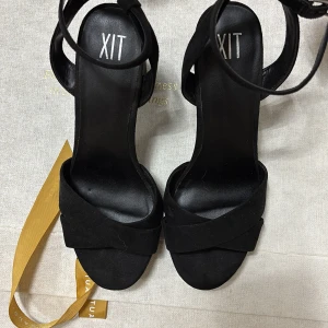Svarta sandaler med klack från XIT - Stilrena svarta klackskor från XIT med breda korslagda remmar framtill och justerbar rem runt vristen. Skorna har öppen tå och en stabil hög blockklack. Ytan är i mockaliknande material som ger en lyxig känsla. Perfekta för att lyfta din outfit.