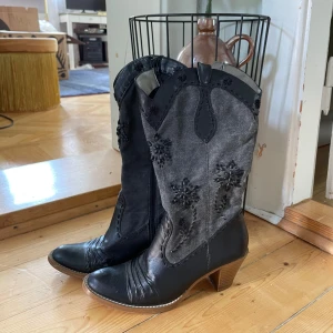 Svarta cowboy boots med broderi - Snygga cowboy boots i svart och mörkgrått med broderade blommor på skaftet. Skorna har klack, rund tå och dragkedja på insidan. Ovandelen är i skinn och textil, med dekorativa sömmar och detaljer. Perfekta för dig som vill sticka ut med westernstil 🖤Storlek 37