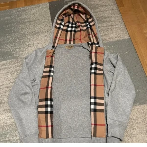 Burberry kofta - Snygg grå hoodie från Burberry med dragkedja och klassiskt rutigt foder i huvan. Tröjan har snörning i huvan, broderad logga på bröstet och mjukt bomullsmaterial. Perfekt för dig som gillar stilrena och exklusiva streetwear-vibbar.