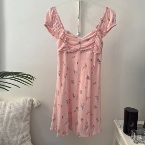 Rosa blommig kortklänning Gina Tricot - Supersöt rosa kortklänning från Gina Tricot med små blommor i olika färger. Klänningen har korta puffärmar, rynkade detaljer vid bysten och en lätt utställd kjol. Perfekt för sommarens alla tillfällen!