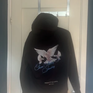 Svart HOODRICH hoodie med duvor - Svart hoodie från HOODRICH med stor vit logga och tryck av två duvor på framsidan SOM ÄR LIMITED EDITION. Baksidan har ett större tryck med tre duvor och blå text. Klassisk huva med snörning och känguruficka framtill. Perfekt för dig som gillar streetwear vibes.
