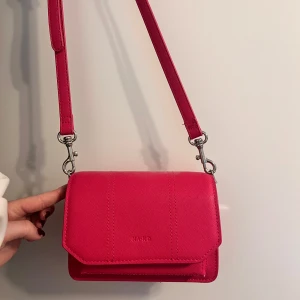 Rosa axelväska från NA-KD - Snygg axelväska från NA-KD i stark rosa färg med stilren, boxig form. Väskan har justerbar axelrem, silverfärgade metalldetaljer och ett huvudfack men även flera olika fack!