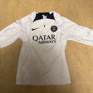 Vit långärmad PSG fotbollströja Nike - Snygg vit långärmad fotbollströja från Nike med Paris Saint-Germain-logga på bröstet och Qatar Airways som sponsor. Tröjan har en svart detalj vid halsen och dragkedja framtill. Perfekt för dig som vill representera PSG på planen eller läktaren.