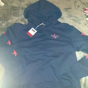 Mörkblå crop hoodie Tommy Jeans strl XXS - Snygg mörkblå hoodie från Tommy Jeans med stora tryckta stjärnor i rött på ärmarna och ett stort logotryck på ryggen. Tröjan har huva med snörning och är tillverkad i mjuk bomull. Perfekt för dig som gillar streetstyle och vill sticka ut. 
