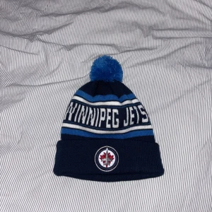 Winnipeg Jets blå stickad mössa - Snygg stickad mössa i mörkblått och ljusblått med vit text 'WINNIPEG JETS' runt om. Mössan har en blå tofs på toppen och broderade loggor fram och bak. Perfekt för dig som vill visa ditt stöd för Winnipeg Jets under kalla dagar. pris diskuterbart