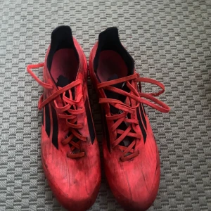 Röda Adidas fotbollsskor med dobbar - Säljer ett par röda Adidas fotbollsskor med svarta detaljer och klassiska tre ränder på sidan. Skorna har snörning och dobbar för bra grepp på planen. Lätta och smidiga, perfekta för dig som vill sticka ut på matchen.