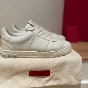Vita Valentino Garavani sneakers - Stilrena vita sneakers från Valentino Garavani i skinn med klassisk rund tå och platt sula. Skorna har diskreta nitar på hälen och vita snören för en clean look. Perfekta för dig som gillar minimalistisk och lyxig stil.