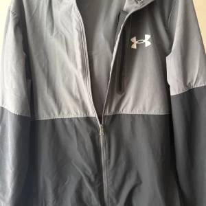 Grå vindjacka från Under Armour - ( Endast att kedjan krånglar till ibland men annars nyskick) Snygg tvåfärgad grå vindjacka från Under Armour med huva och dragkedja framtill. Jackan har en sportig look, diskret logga på bröstet och en bröstficka med dragkedja. Perfekt för dig som gillar stilren och funktionell design. 