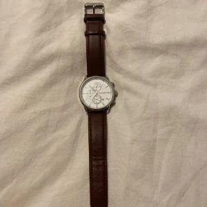 Studsgaard klocka | brun läderrand  - Stilren klocka från Studsgaard med vit urtavla, silverfärgat boett och flera små urtavlor. Remmen är i äkta brunt läder och boetten är i rostfritt stål. Snygg och klassisk design som passar till många outfits.