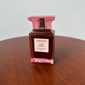 Tom Ford Lost Cherry 100ml parfym - Tom Ford Lost Cherry Eau de Parfum 100 ml i en lyxig, fyrkantig glasflaska med röd och rosa ton. Flaskan har en elegant, genomskinlig rosa kork och etikett framtill. Doften är känd för sina söta och fruktiga körsbärsnoter och exklusiva känsla.