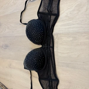 Svart mesh korsett från Censored - Snygg svart korsett från Censored med meshdetaljer och glittrande små paljetter över kuporna. Axelbandslös design som ger en cool och festlig vibe. Perfekt att styla under en kavaj eller som statement-plagg till din outfit.