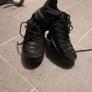 Svarta Nike Vapormax sneakers - Svarta Nike Vapormax sneakers med cool, futuristisk design och synlig luftbubbelsula. Skorna har snörning och är tillverkade i syntetmaterial och mesh för en sportig vibe. Perfekta för dig som gillar streetstyle och vill ha något som sticker ut.