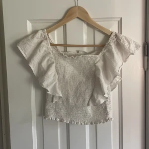 Vit volangtopp från Kappahl - Supersöt vit topp från Kappahl med smockad kropp och stora volangärmar. Toppen är croppad och har en lätt rynkad kant nertill. Perfekt för dig som gillar romantisk och luftig stil. Tillverkad i mjuk bomull som känns skön mot huden. Den är i storlek 158/164 men skulle säga att den passar xs-m ungefär då den är stretchig. Köpt för 199 kr. 