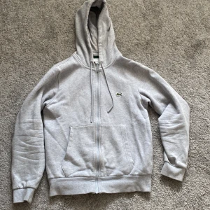 Grå hoodie från Lacoste med dragkedja - Snygg ljusgrå hoodie från Lacoste i klassisk modell med dragkedja, huva med snörning och känguruficka. Broderad krokodil-logga på bröstet. Mjuk insida och ribbade muddar. Perfekt för chill dagar eller när du vill ha en clean streetstyle. Har haft ett litet hål på baksidan fast som knappt syns då det har blivit sytt. Pris kan diskuteras!