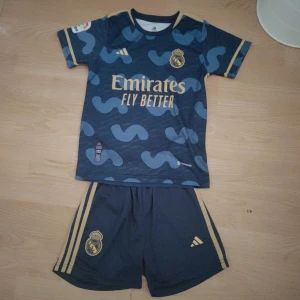 Real Madrid fotbollströja och shorts - Snyggt Real Madrid matchställ från Adidas med kortärmad tröja och shorts. Tröjan är marinblå med guldiga detaljer och tryck, samt ett unikt blått mönster. Materialet är lätt och andas, perfekt för fotbollsträning eller match. Shorts med guldiga ränder och klubbmärke. Storlek 152 och den har vini Jr på ryggen men nummer tjugo på den 