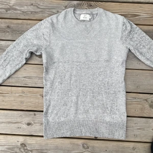 Grå stickad tröja från Emilio - Snygg ljusgrå stickad tröja från Emilio med rund halsringning och ribbade muddar. Enkel och stilren design som passar perfekt till jeans eller chinos. Mjuk och skön känsla, perfekt för kyligare dagar. Har dock ett litet hål i högra hörnet!