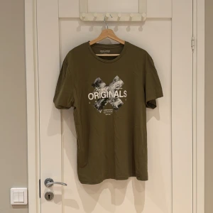 Olivgrön t-shirt Jack & Jones Originals - Olivgrön t-shirt från Jack & Jones Originals med coolt svartvitt tryck och text på bröstet. Klassisk rund hals och normal passform. Tillverkad i mjuk bomull, perfekt för en avslappnad stil.