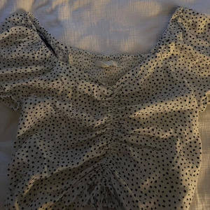 Prickig blus med puffärm och snörning - Supersöt vit blus med svarta prickar, puffiga korta ärmar och smockad rygg. Toppen har liten v-ringning och snygg snörning framtill som ger en rynkad effekt. Perfekt för dig som gillar romantisk och trendig stil.