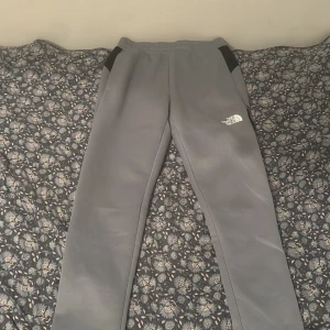 Grå mjukisbyxor från The North Face - Snygga grå mjukisbyxor från The North Face med svarta detaljer vid fickorna och logga på låret. Byxorna har elastisk midja och raka ben, perfekta för chill eller träning. Sköna och stilrena, passar till det mesta.
