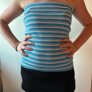 Randig bandeau tubtopp från Mango - Säljer en ribbad bandeau tubtopp från Mango i blått med vita och rosa ränder. Toppen är ärmlös och sitter tight mot kroppen, perfekt för varma dagar. Superenkel att styla med både jeans, shorts och kjol för en chill look. Passar även som tubkjol. 