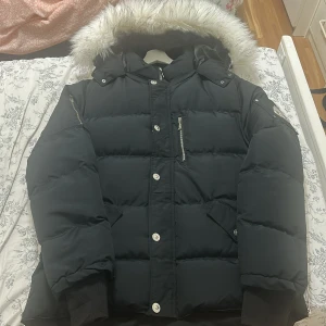 Svart dunjacka med fuskpäls från Down Filled storlek xxl - Snygg svart dunjacka från Down Filled med fluffig vit fuskpäls på huvan. Jackan har dragkedja, knappar och flera fickor, inklusive en med dragkedja på bröstet. Perfekt för kalla vinterdagar och har en riktigt maffig, puffer-look.