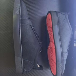 Svarta sneakers från Christian Louboutin - Snygga svarta sneakers från Christian Louboutin med klassisk röd sula och diskret logga undertill. Skorna har rund tå, snörning och är tillverkade i mocka med svarta detaljer. Perfekta för dig som vill ha en clean och exklusiv look.