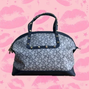 Blå vintage DKNY handväska med nitar - Snygg vintage handväska från DKNY i blått och grått med logomönster över hela väskan. Väskan har både handtag och justerbar avtagbar axelrem i blått skinn, dekorerad med coola guldfärgade nitar. Stängs med dragkedja och har rymligt svart foder inuti. Super söta vintage y2k.      Ungefärliga mått: Längd nedtill: 31 cm Topp till botten exklusive handtag: 25 cm Topp till botten inklusive handtag: 35 cm Sida till sida nedtill: 15 cm Sida till sida i mitten: 25 cm