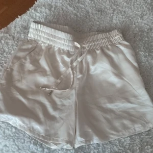 Vita satinshorts med snörning ny - Snygga vita shorts i glansigt satinmaterial med bred resår i midjan och snörning framtill. De har en avslappnad passform och är perfekta för varma dagar. Shortsen har fickor på sidorna och en mjuk känsla mot huden. Passar xs . Endast provade 
