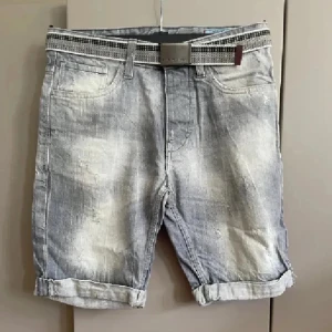 Grå jeansshorts med bälte - Snygga grå jeansshorts med coolt tvättad look och uppvikta benslut. Shortsen har fem klassiska fickor och medföljer ett grått bälte med metallspänne. Perfekta för en avslappnad stil under varma dagar.