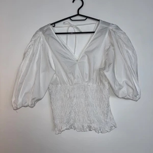 Vit blus med puffärmar och smock - Superfin vit blus med djup v-ringning, puffiga trekvartsärmar och smockad midja. Blusen har knytband i nacken och volangkant nedtill för en extra söt touch. 