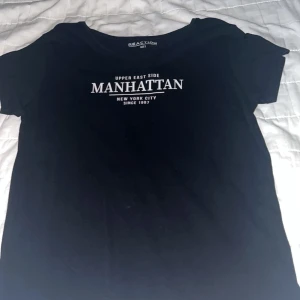 Svart Manhattan t-shirt från Reaction - Svart t-shirt från Reaction med vit text på bröstet där det står 'Upper East Side Manhattan New York City'. Klassisk rund halsringning och korta ärmar. Skön och enkel modell i mjukt bomullstyg, perfekt till jeans eller shorts.