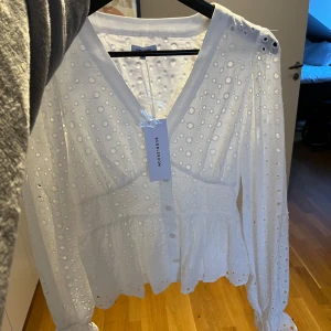Vit Anglaise Blouse - från Bubbleroom - Superfin vit blus från Bubbleroom med broderade hål och v-ringning. Blusen har långa ärmar, knappar framtill och lätt volang nertill. Oanvänd. 