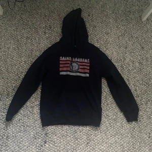  Saint Laurent hoodie med tryck - Marinblå hoodie från Saint Laurent med huva. Framsidan har ett tryck med röda och vita ränder samt logga och text. Långärmad och i mjukt material, perfekt för en chill och snygg look. Vet ej om den är äkta därför billigt pris.