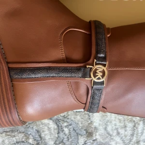 Bruna knähöga stövlar från Michael Kors - Snygga bruna knähöga stövlar från Michael Kors med guldfärgad MK-detalj och mörkbruna remmar runt ankeln. Dragkedja på sidan och rund tå. Perfekta för att lyfta din höst- eller vinterstil.