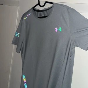 Grå t-shirt från Under Armour - Säljer en grå t-shirt från Under Armour med färgglada loggor och textdetaljer på bröstet, ärmen och nedtill. T-shirten har rund halsringning och är tillverkad i ett syntetmaterial som passar perfekt till träning.
