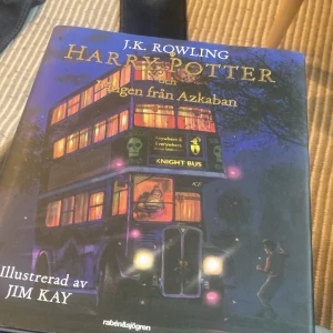 Harry Potter och fången från Azkaban - Illustrerad utgåva av 'Harry Potter och fången från Azkaban' av J.K. Rowling, med färgstarka illustrationer av Jim Kay. Omslaget visar den ikoniska riddarbussen i lila och gult mot en mörk bakgrund. Boken är på svenska och utgiven av Rabén & Sjögren.